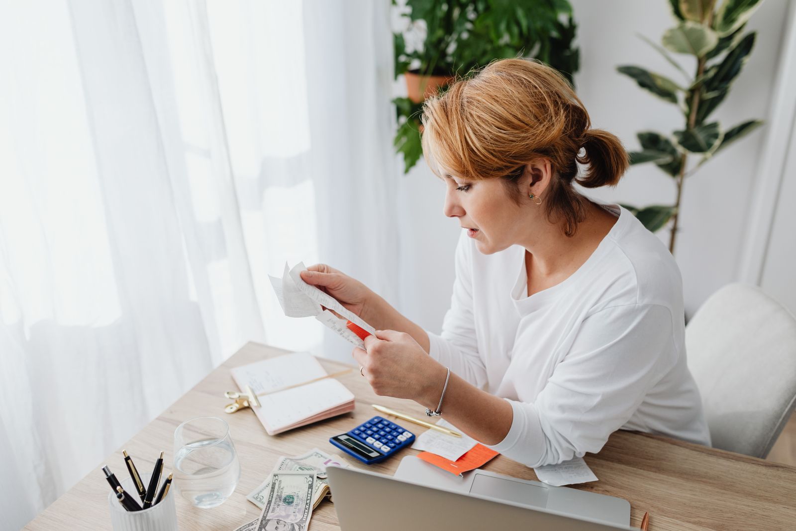 A woman at a bright desk sorting receipts beside a laptop